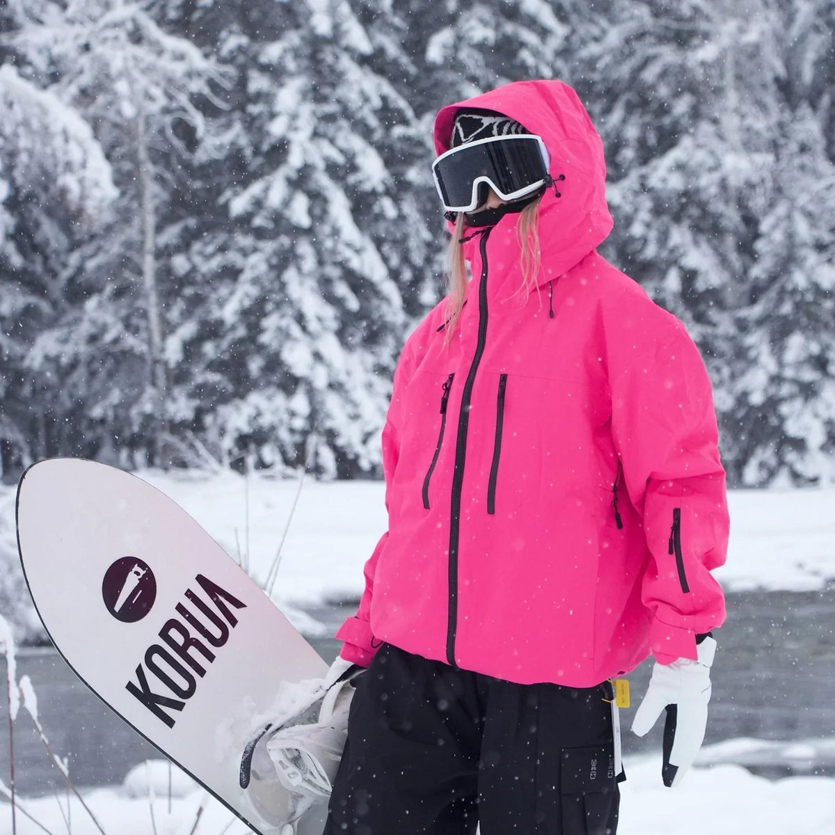 Model in a bright pink hooded jacket with black zips, carrying a snowboard in a snowy forest. 5