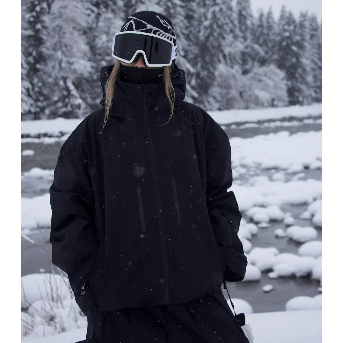 Model wearing the black version of the oversized hooded jacket, hands in pockets, beside a snowy river. 2