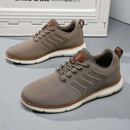 Pair of taupe knitted mesh trainers with cream soles and tan leather trim, standing on a grey floor in front of a storage box. 2