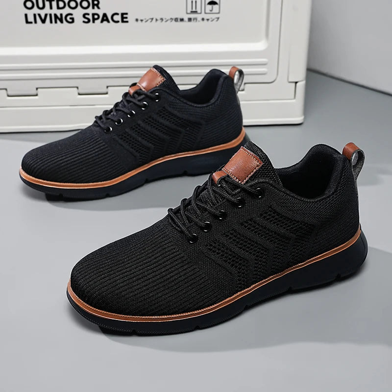 Pair of all-black knitted mesh trainers with matching dark soles and tan piping, shown on a grey floor beside a storage box. 3