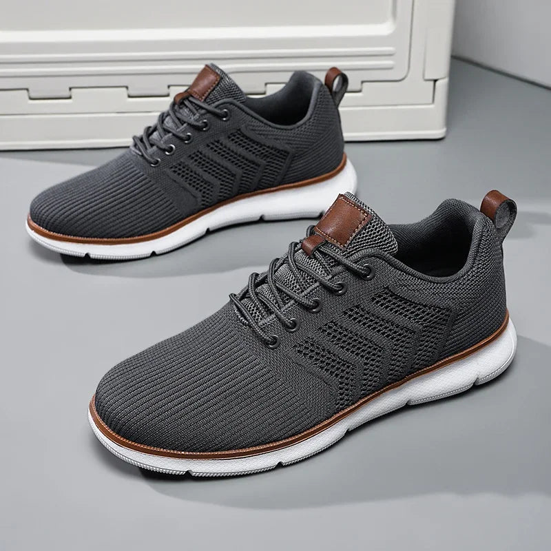 Pair of dark grey knitted mesh trainers with white soles and tan leather trim, displayed on a grey floor. 4
