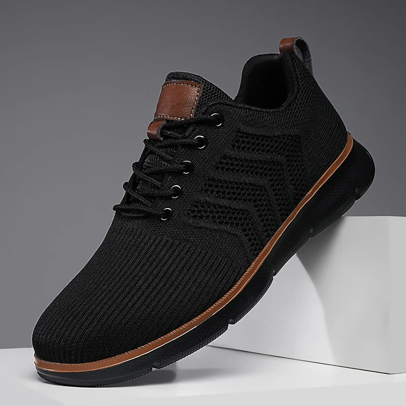Single all-black mesh trainer with tan piping, angled on a white block to show the laces and ventilated side panel. 6
