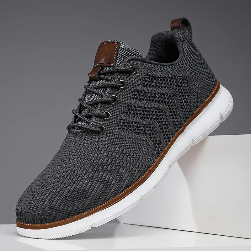 Single dark grey mesh trainer with white sole and tan trim, angled on a white block to show the laces and perforated panel. 7