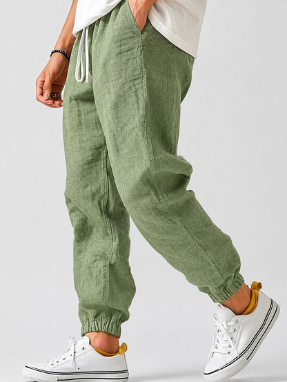 Sage green jogger trousers on a model walking, with a white drawstring, side pocket and gathered cuffs above white trainers. 6