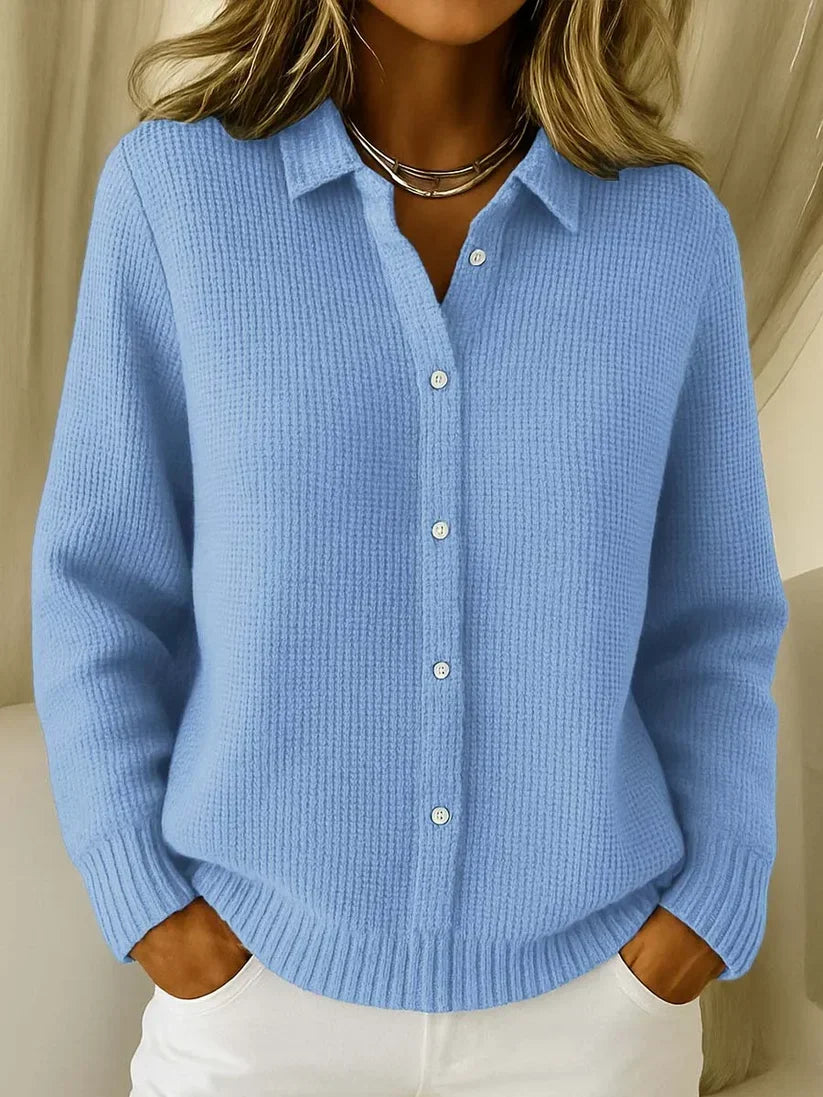 Model wearing the waffle-knit collared jumper in light blue, buttoned down the front, with hands in the pockets of white trousers. 1