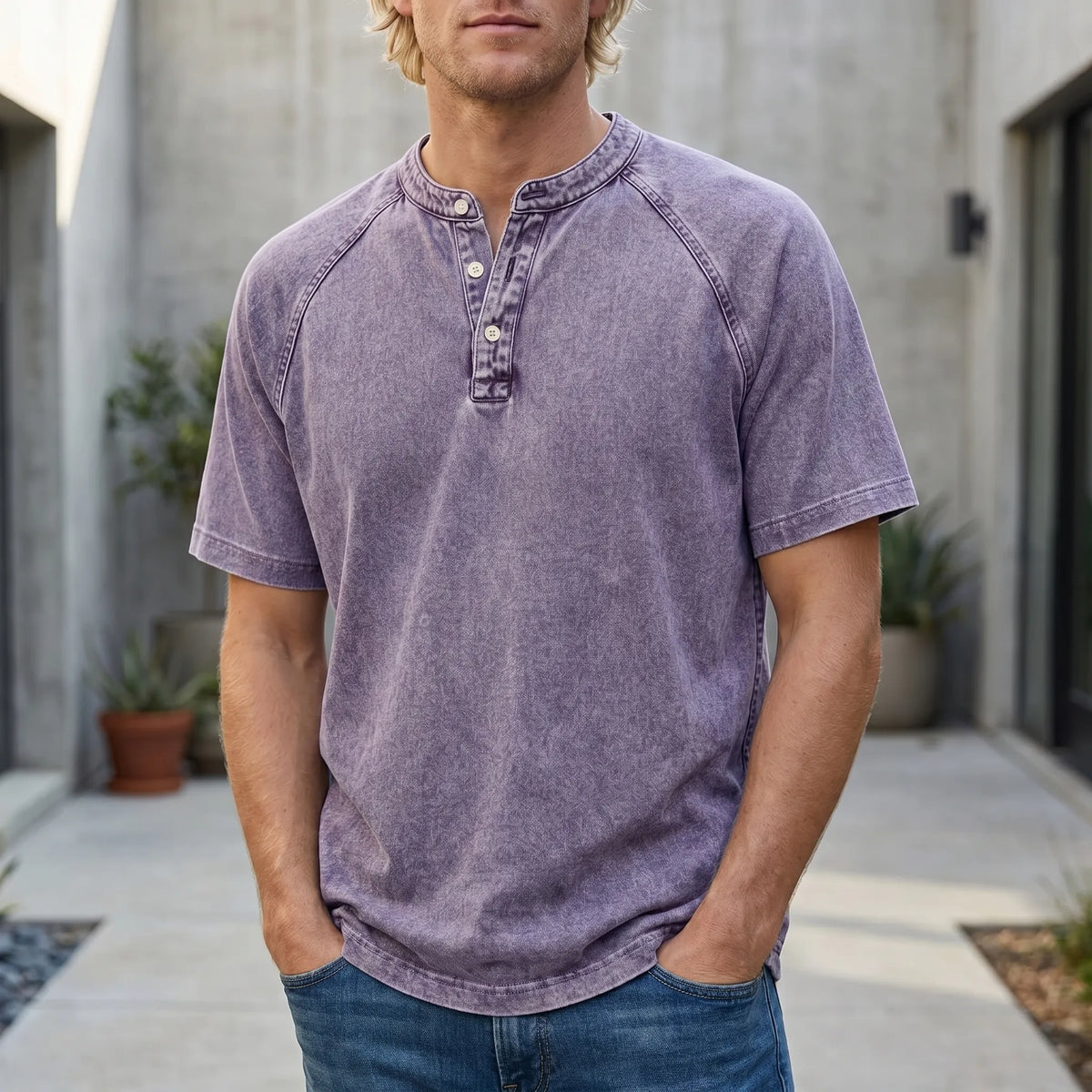 Fair-haired man in a faded purple raglan-sleeve Henley with hands in jeans pockets, standing outdoors in a concrete courtyard. 1