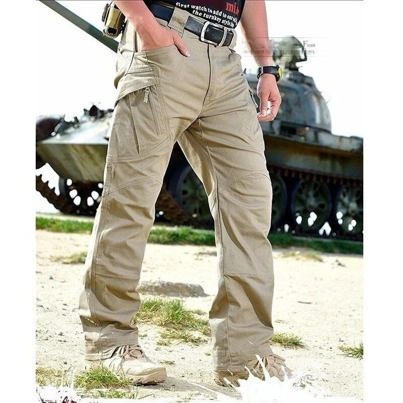 Model in khaki cargo trousers with a belt and desert boots, one hand in a pocket, standing on grass in front of a tank. 9