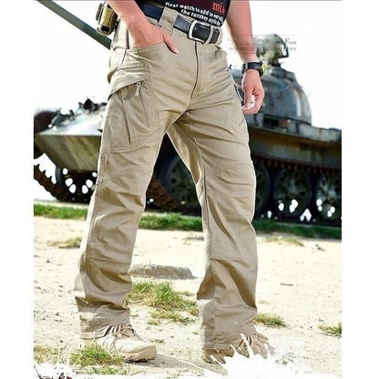 Model in khaki cargo trousers with a belt and desert boots, one hand in a pocket, standing on grass in front of a tank. 9