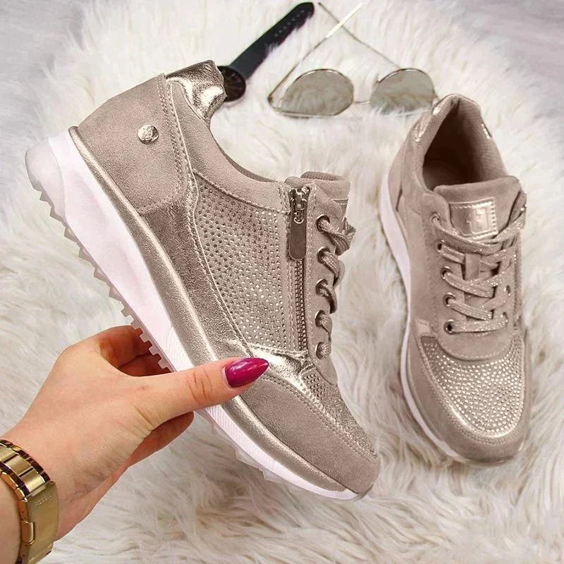 Beige-gold trainers held in one hand over a fluffy white rug, showing the chunky white sole, zip and lace-up front. 7
