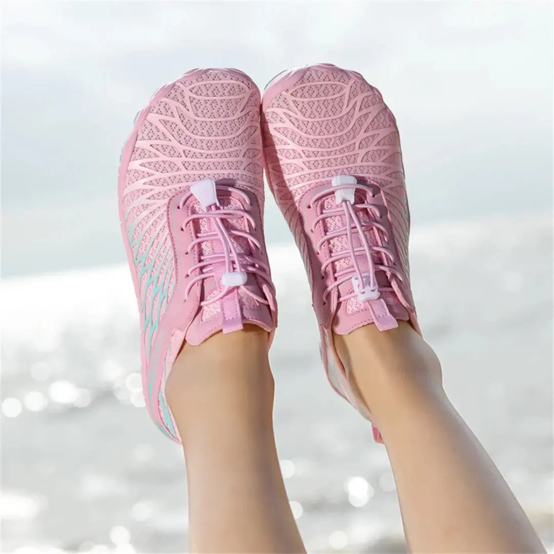 Pink mesh barefoot shoes seen from above on raised feet, showing the toggle laces and wide toe area, with sparkling water behind. 14