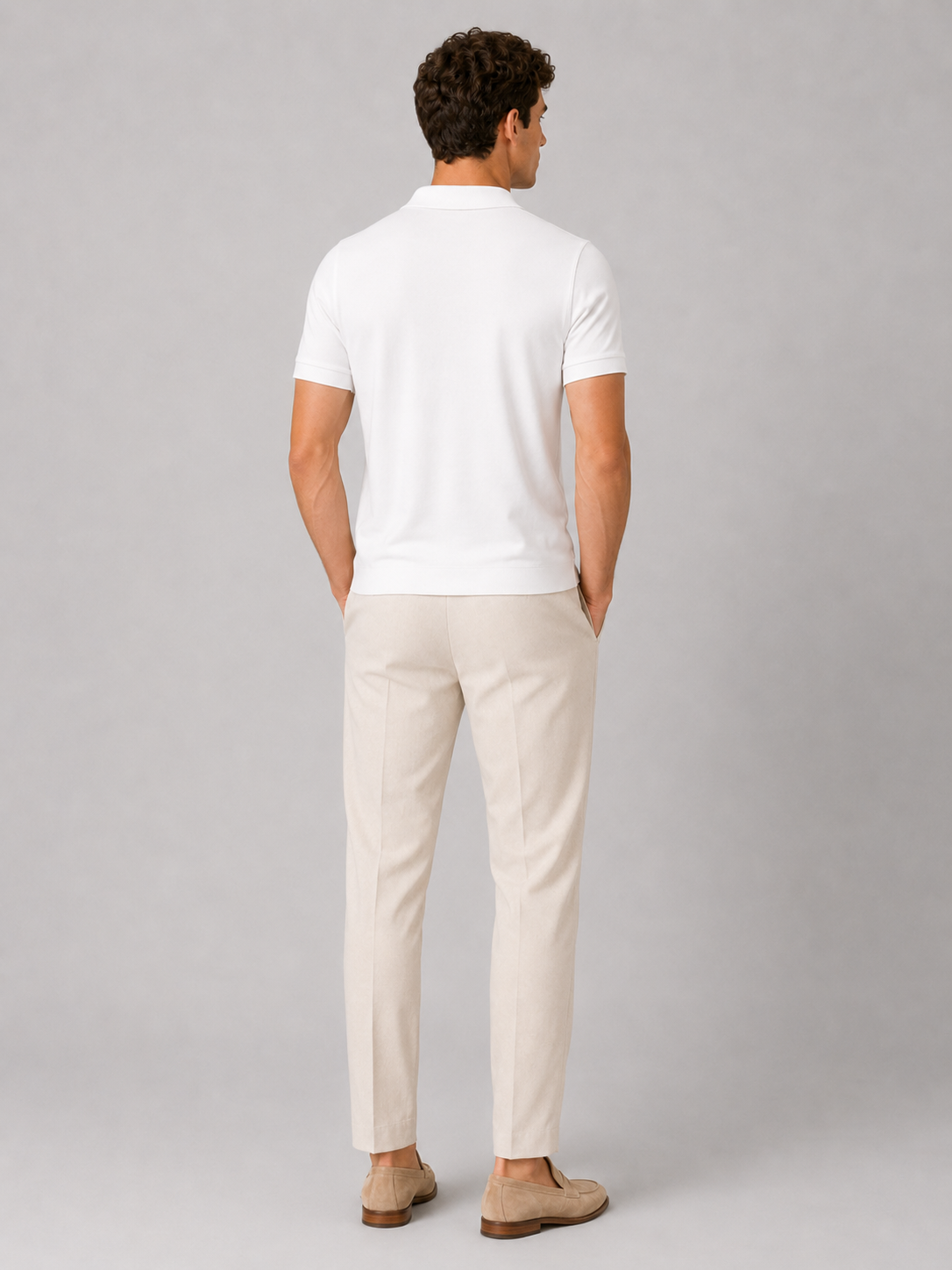 Back view of the white polo on a standing model, showing the plain back, short sleeves and ribbed hem, with cream trousers and loafers. 4