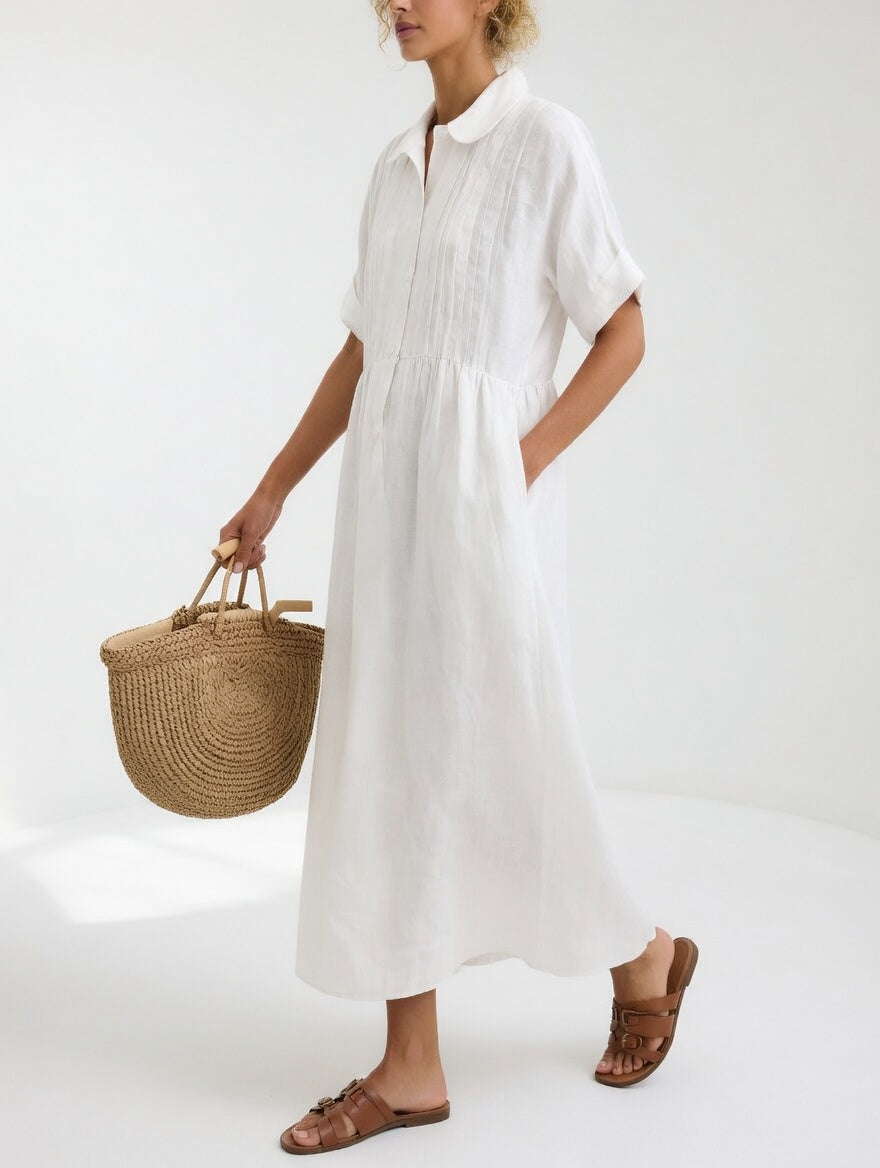 Model in the white dress seen from the side, one hand in a pocket, carrying a woven straw tote and tan sandals. 1
