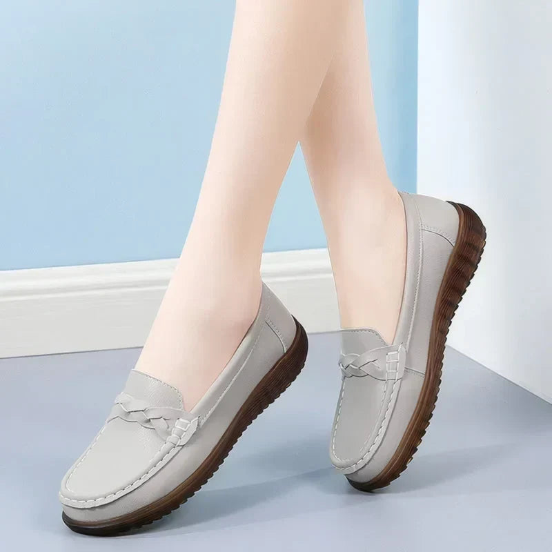Light grey loafers with braided strap, one heel lifted to show the dark ridged sole, against a blue wall. 12