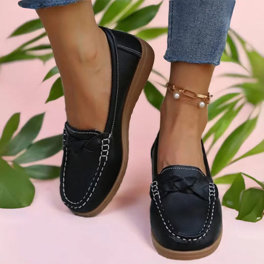 Black loafers with plaited strap and white stitching worn with cropped jeans and a pearl anklet against pink with green leaves. 2