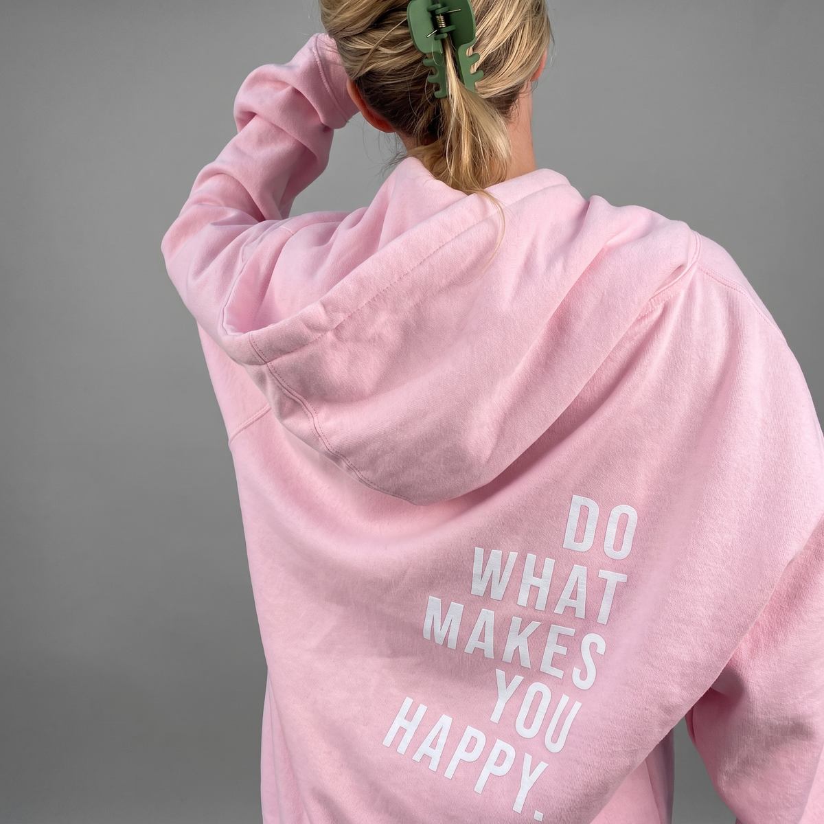 Model seen from behind in a light pink hoodie with the hood down and a large white slogan print on the back. 1