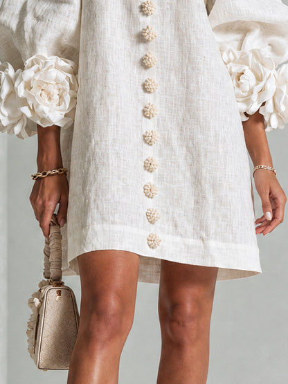 Close-up of the cream dress hem and lower front, with flower buttons, straight hem and rose cuffs beside a small handbag. 6