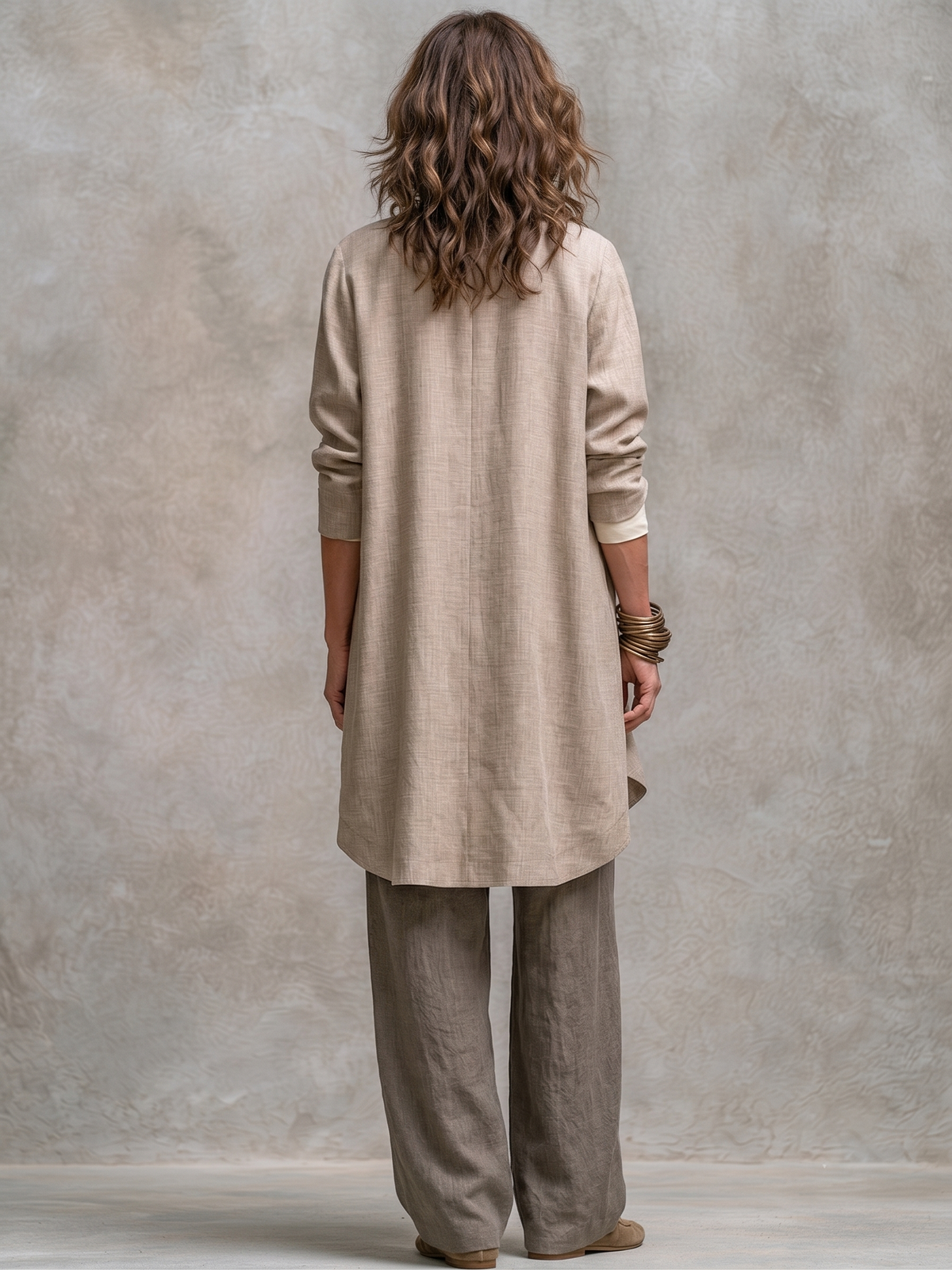 Back view of the long beige jacket with its centre back seam and curved hem, worn over taupe wide-leg trousers. 6