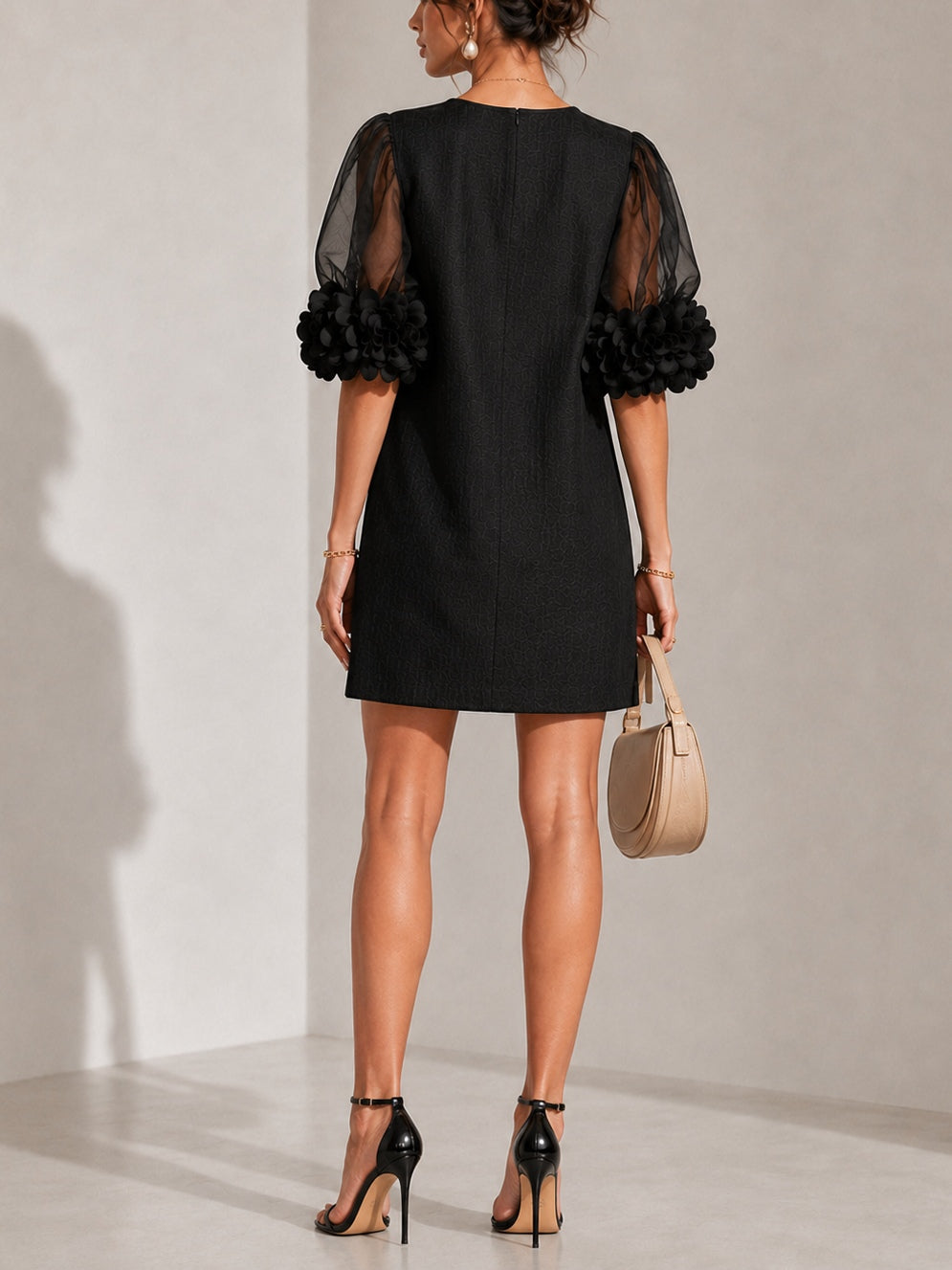 Back view of the black dress on a model, showing the centre seam, sheer puffed sleeves and floral cuffs from behind. 3