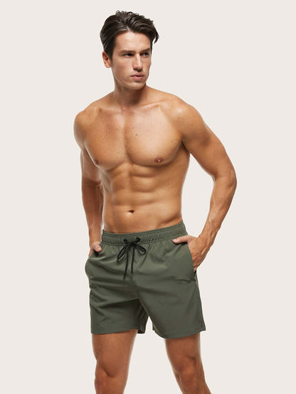 Model standing with both hands in the pockets of olive green swim shorts, seen from the front at an angle. 5