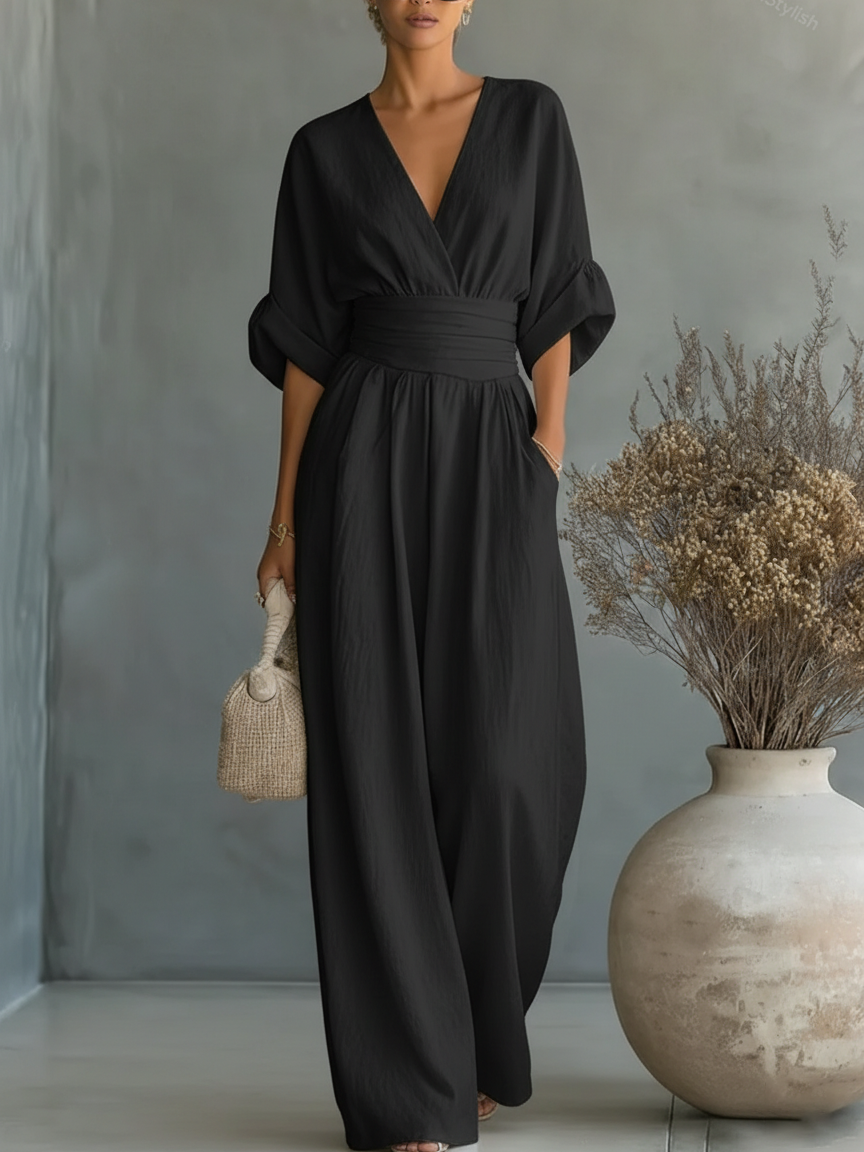 Black jumpsuit on a model, showing the V-neckline, cuffed sleeves and flowing wide legs beside a vase of dried flowers 6