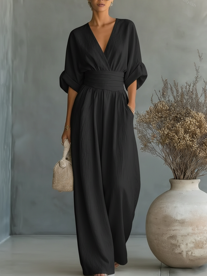 Black jumpsuit on a model, showing the V-neckline, cuffed sleeves and flowing wide legs beside a vase of dried flowers 6
