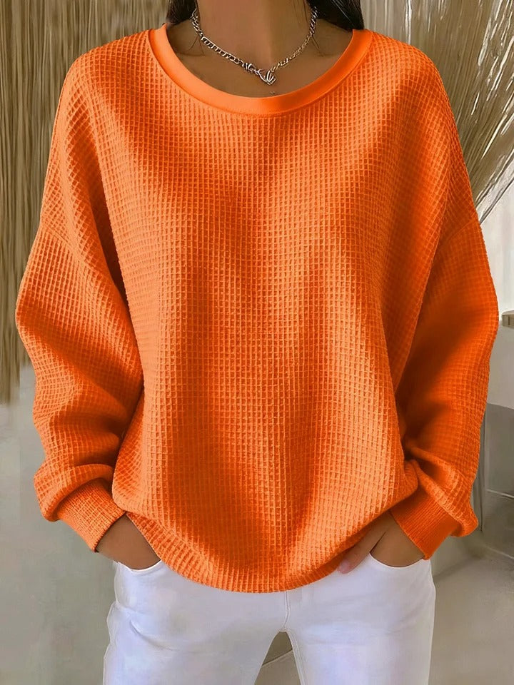 Model wearing the waffle-knit jumper in bright orange, hands in trouser pockets, showing the square textured weave. 8