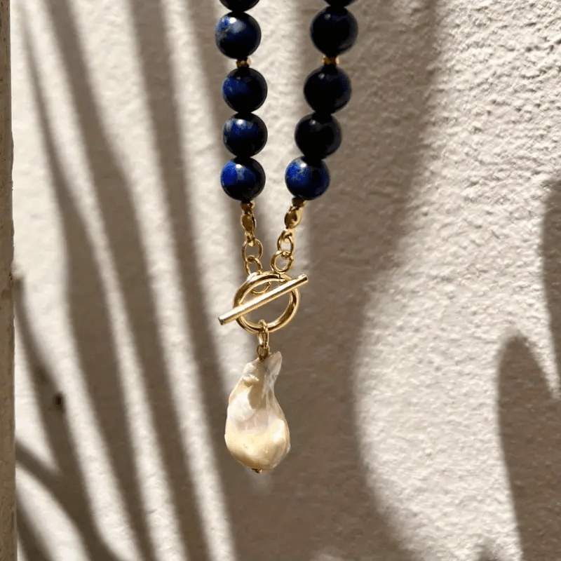 Necklace hanging against a textured white wall with leaf shadows, showing the toggle clasp and pearl 8
