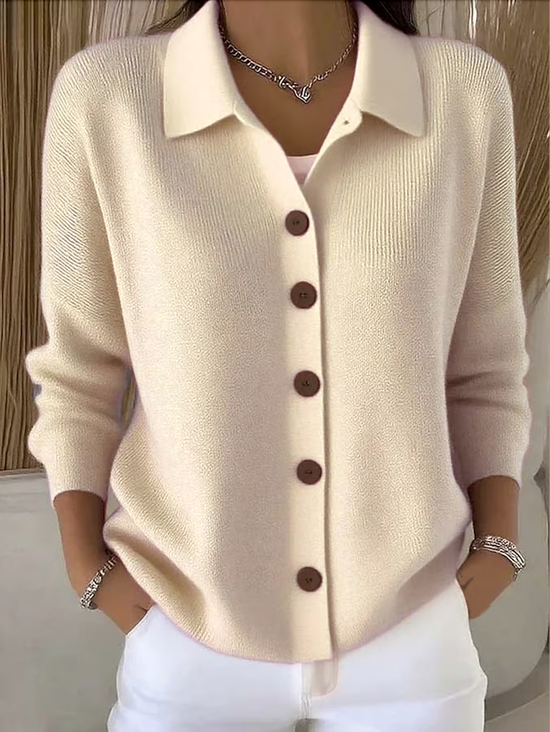 Model wearing the cream knit cardigan with brown buttons and a turned-down collar, hands in pockets, styled with white trousers. 1