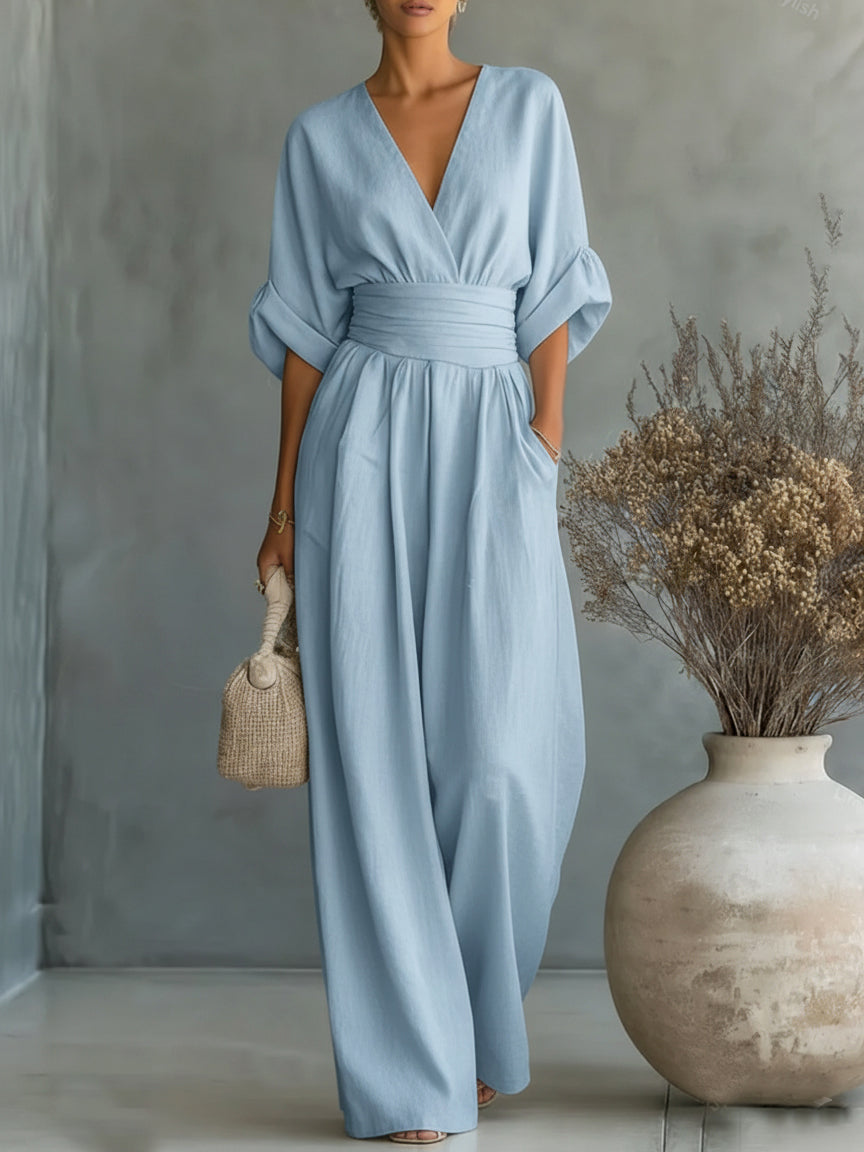 Model wearing the pale blue jumpsuit with a wrapped waist band, holding a woven bag next to a vase 2