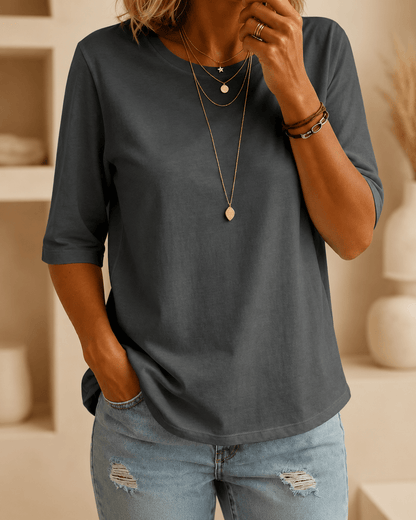 Woman wearing a dark grey crew-neck tee with elbow-length sleeves, layered gold necklaces and ripped light-wash jeans, one hand in her pocket. 1