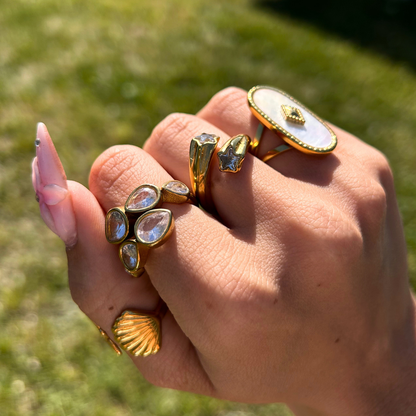 Close-up of a hand outdoors on grass, wearing the pear-crystal ring stacked with gold star, shell and oval mother-of-pearl style rings. 4