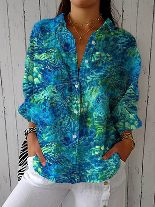 Model wearing the long-sleeved button-up blouse in a blue and green peacock feather print, worn with white trousers and hands in pockets. 1