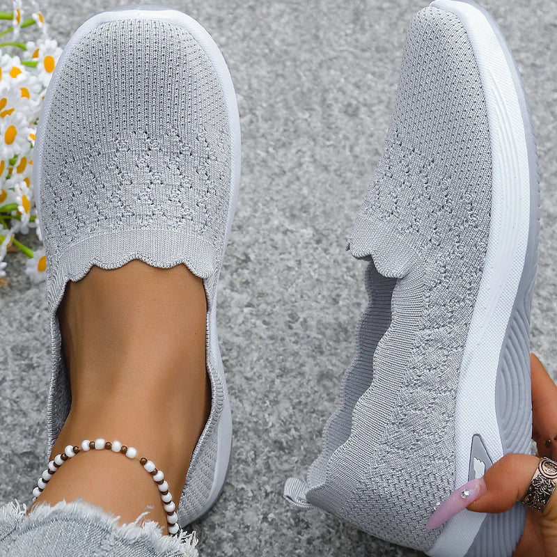 Top-down view of grey knit slip-on shoes with scalloped collar, one worn and one held 11