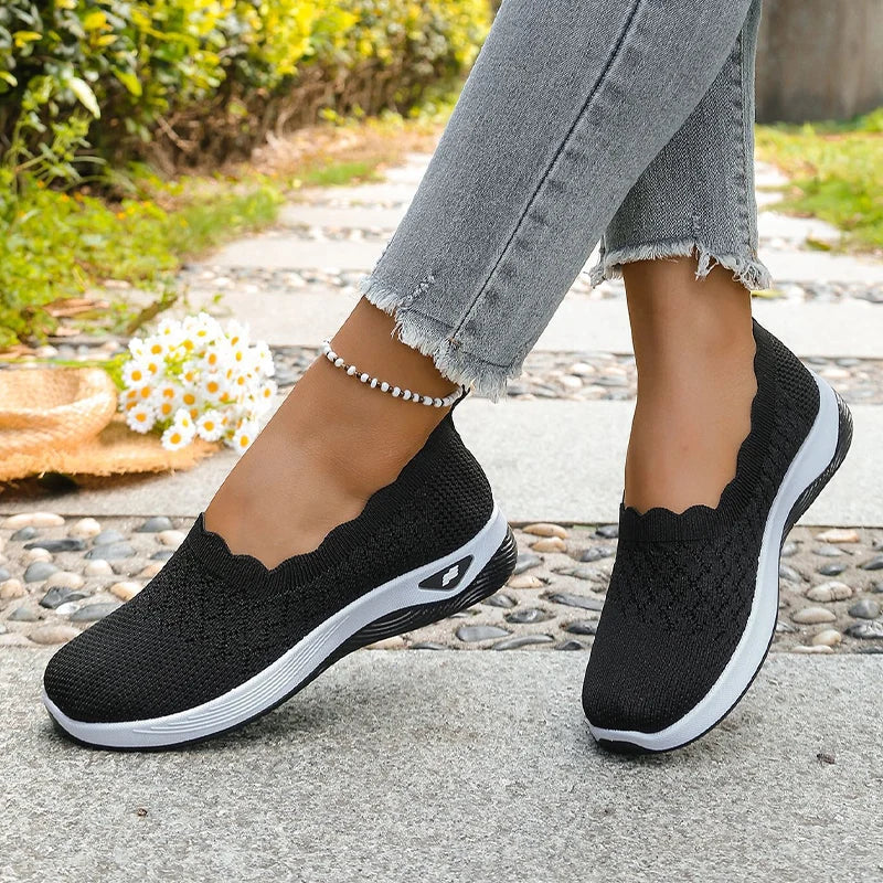 Black knit slip-on shoes with scalloped collar worn mid-step on a paved path 14