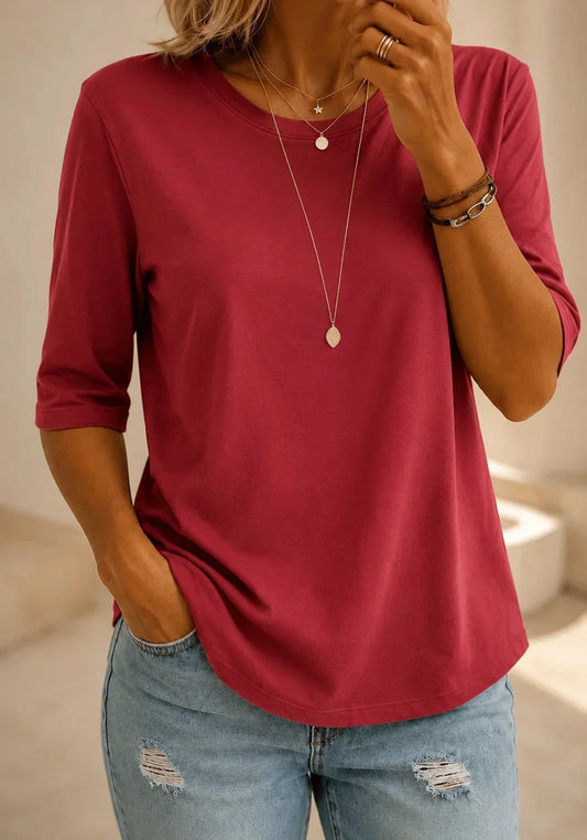 Woman wearing a plain raspberry-red half-sleeve top with a round neck, layered gold necklaces and distressed light-wash jeans, one hand in her pocket. 1