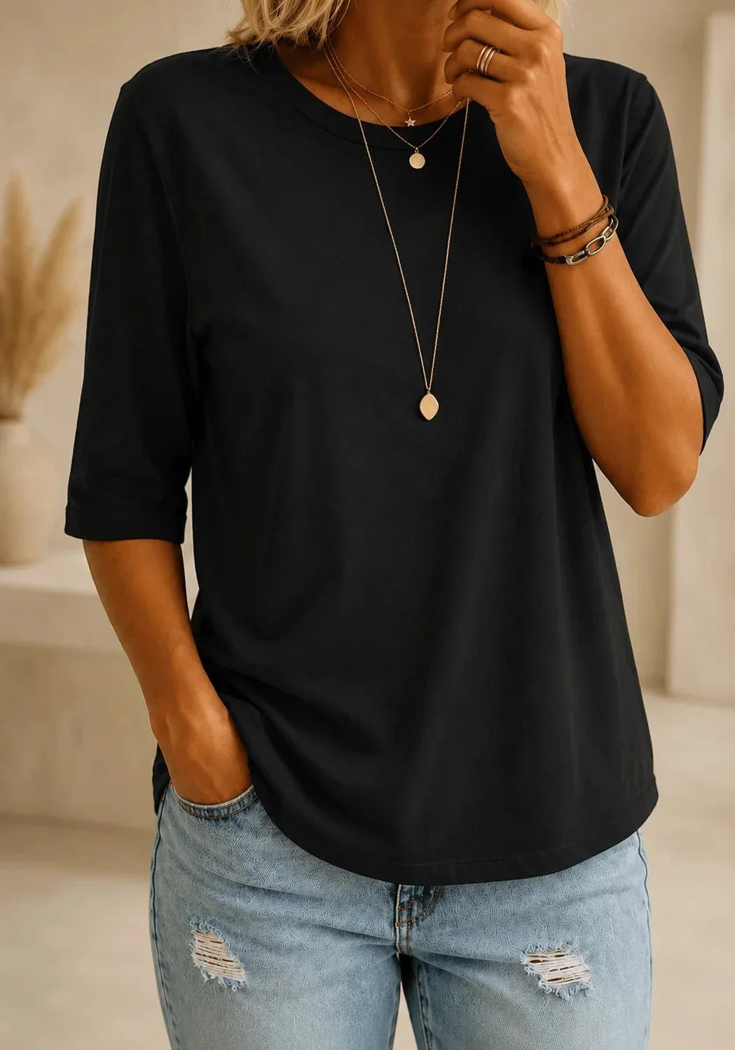Woman wearing a plain black round-neck half-sleeve tee with a curved hem, styled with layered gold necklaces and light distressed jeans. 1