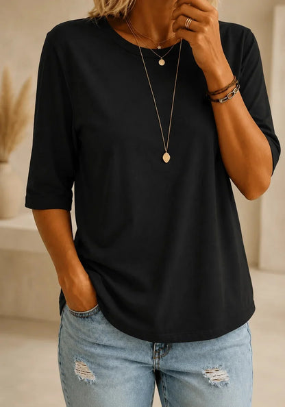 Woman wearing a plain black round-neck half-sleeve tee with a curved hem, styled with layered gold necklaces and light distressed jeans. 1