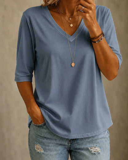 Model wearing a slate blue V-neck top with elbow-length sleeves, layered gold necklaces and distressed light denim jeans, shown from the front. 1