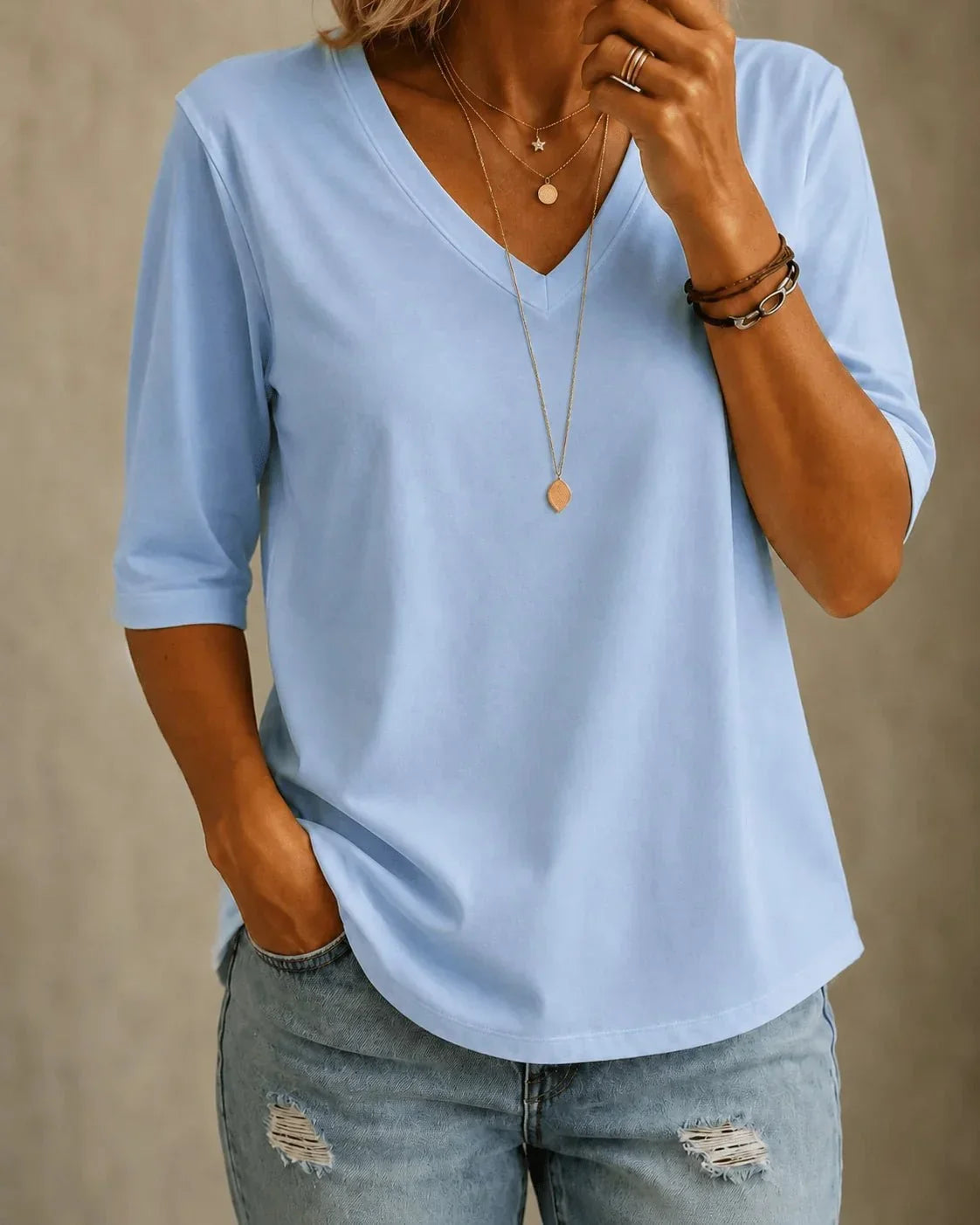 Woman wearing a light blue V-neck tee with elbow-length sleeves, layered gold necklaces and ripped light-wash jeans, one hand in her pocket. 1