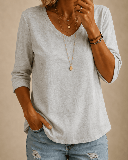 Woman wearing the light grey V-neck tee with layered gold necklaces and distressed light-wash jeans, one hand in pocket 1