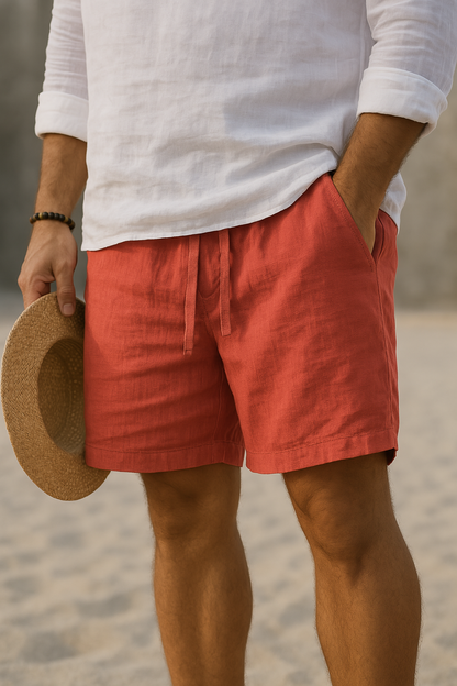 Drawstring shorts in coral red worn by a model on pale sand with a white linen shirt, hand in pocket, straw hat in hand. 7