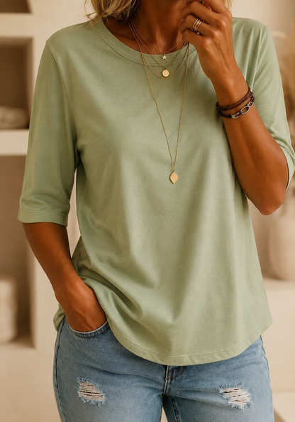 Model wearing the tee in soft sage green with gold pendants and ripped jeans, in a warm interior with shelving behind. 11