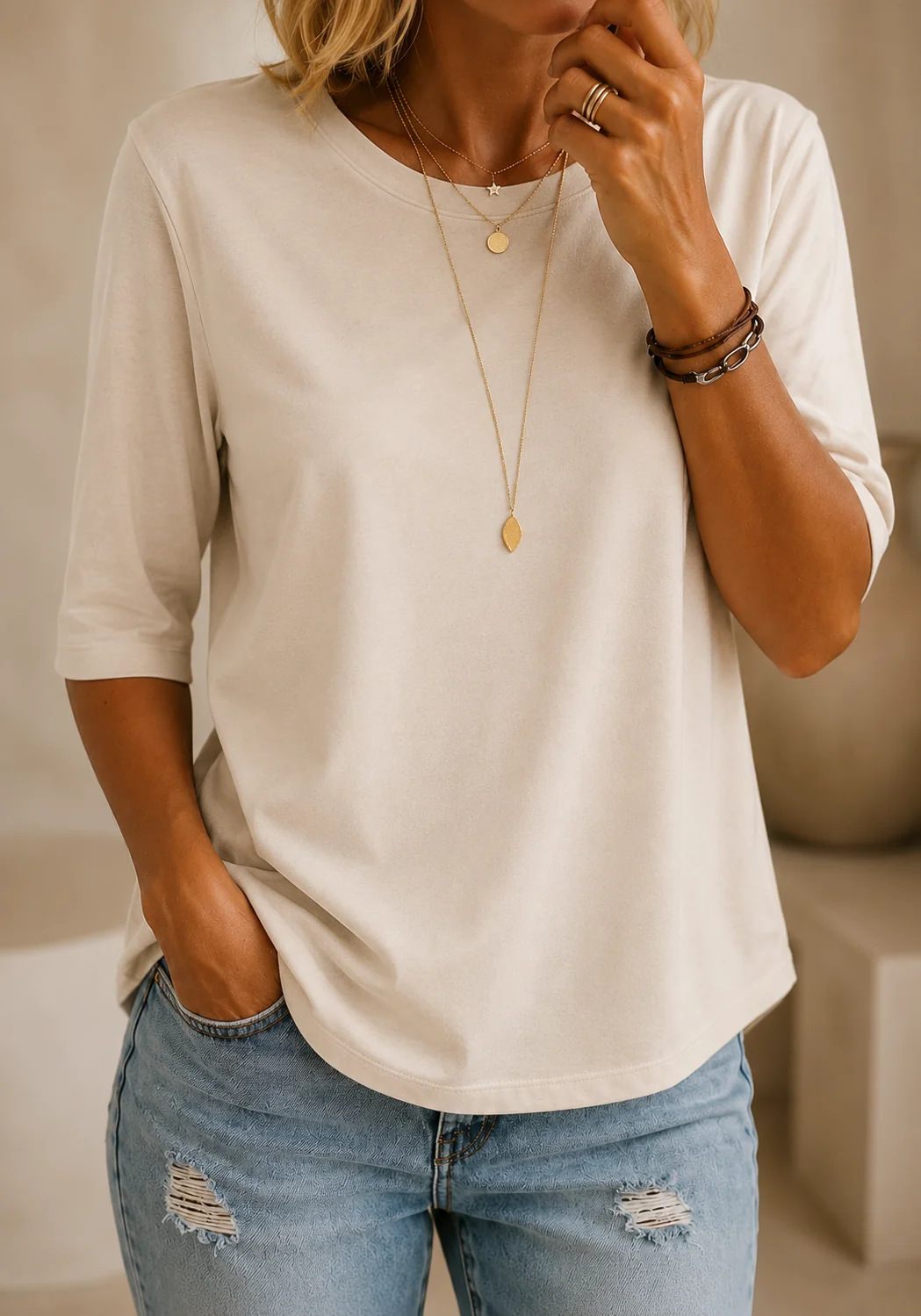 Model wearing the tee in cream with layered gold necklaces and ripped jeans, in a softly lit neutral interior. 10