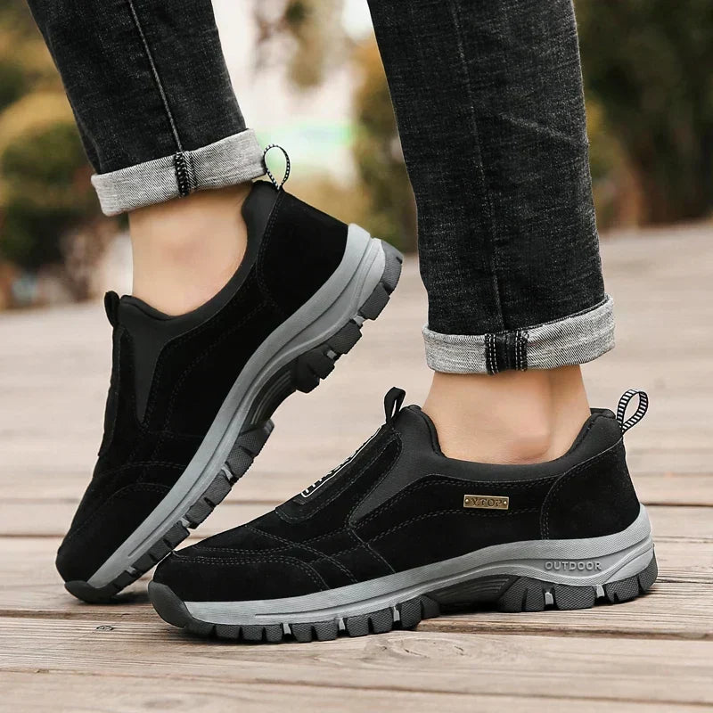 Black slip-on shoes worn by a model in turned-up dark jeans, standing on wooden decking, showing the grey chunky sole. 4