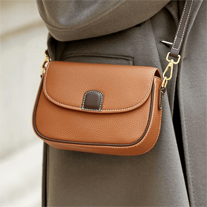 Tan leather crossbody bag with dark brown tab and gold clips, worn against a grey coat 2