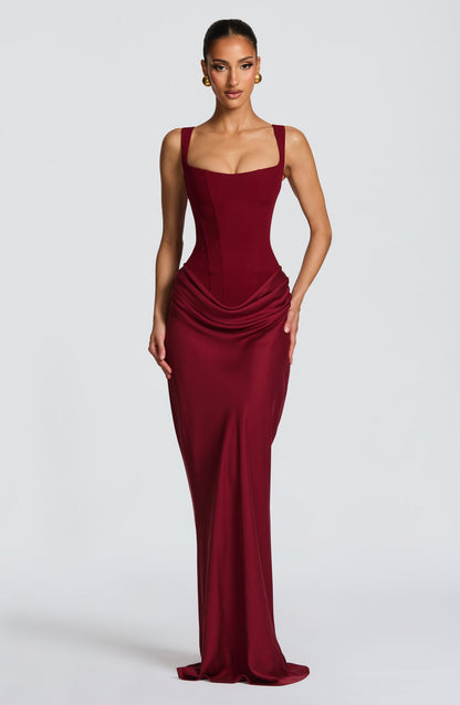 Model wearing the burgundy maxi dress, front view, with square neckline, boned corset bodice and draped hip detail, hand on hip. 22