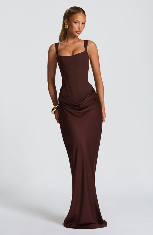 Model facing the camera in the brown corset maxi dress with draped hip detail, gold earrings and a chunky gold bangle. 1