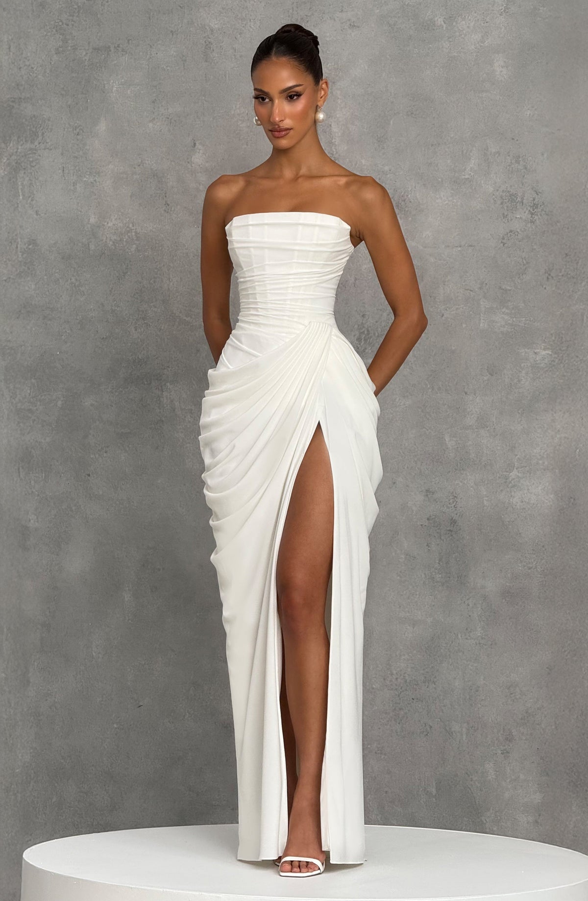 White strapless maxi dress on a white plinth against a grey wall, showing the draped skirt and high slit. 14