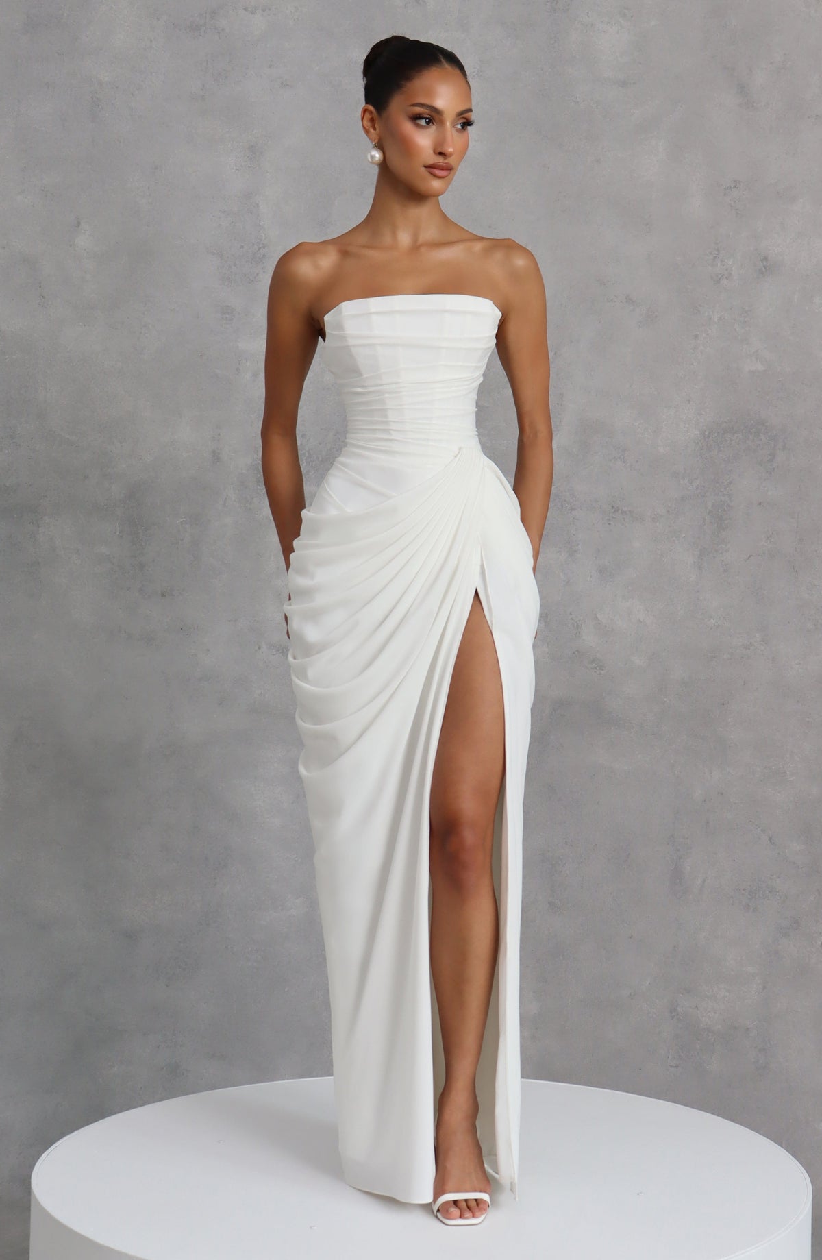 White strapless corset maxi dress styled with pearl earrings against a grey textured backdrop, model looking to the side. 13