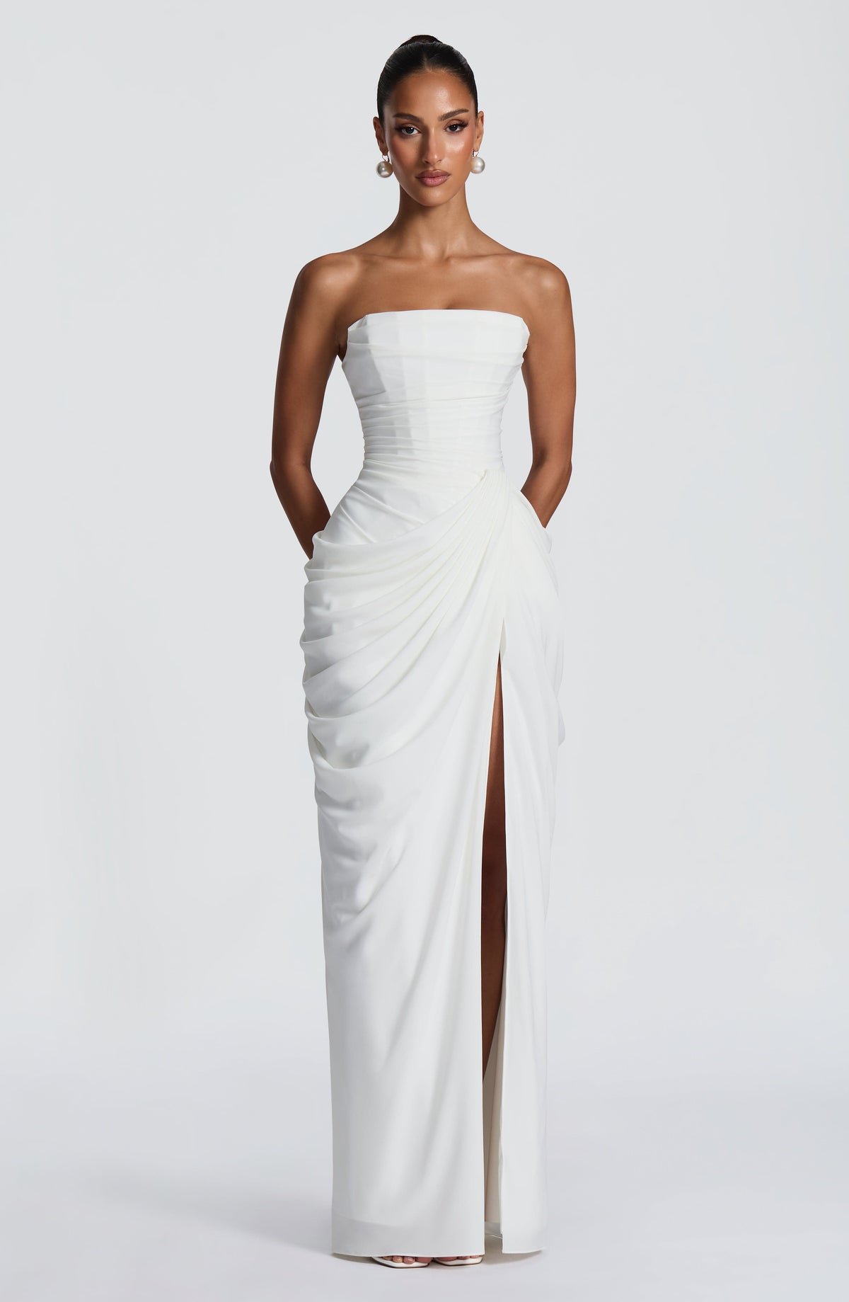 Full-length front view of the white corset maxi dress with high slit, hands in pockets and pearl earrings. 11